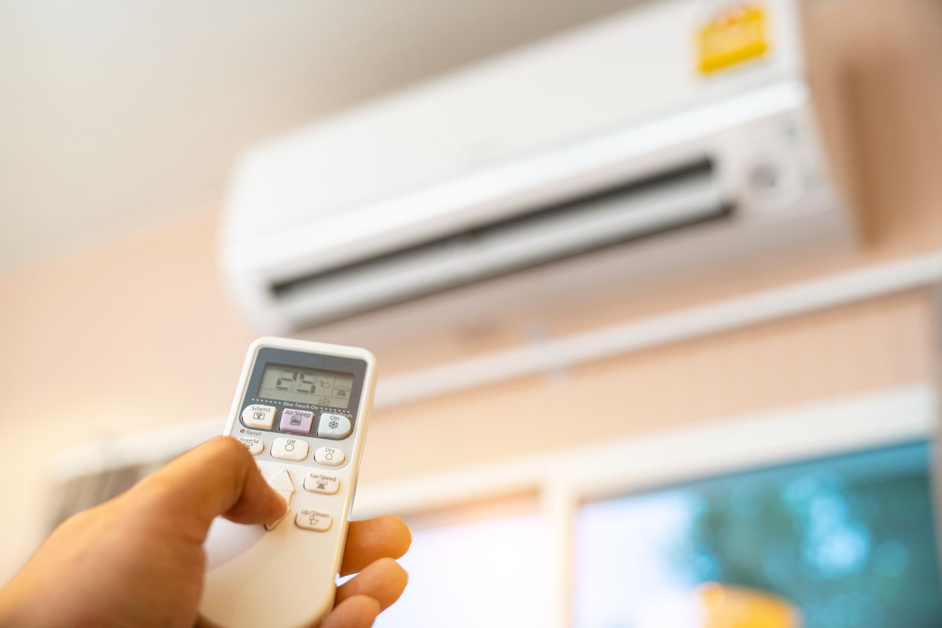 Hand holding AC remote, pointing it at a white wall-mounted air conditioner.