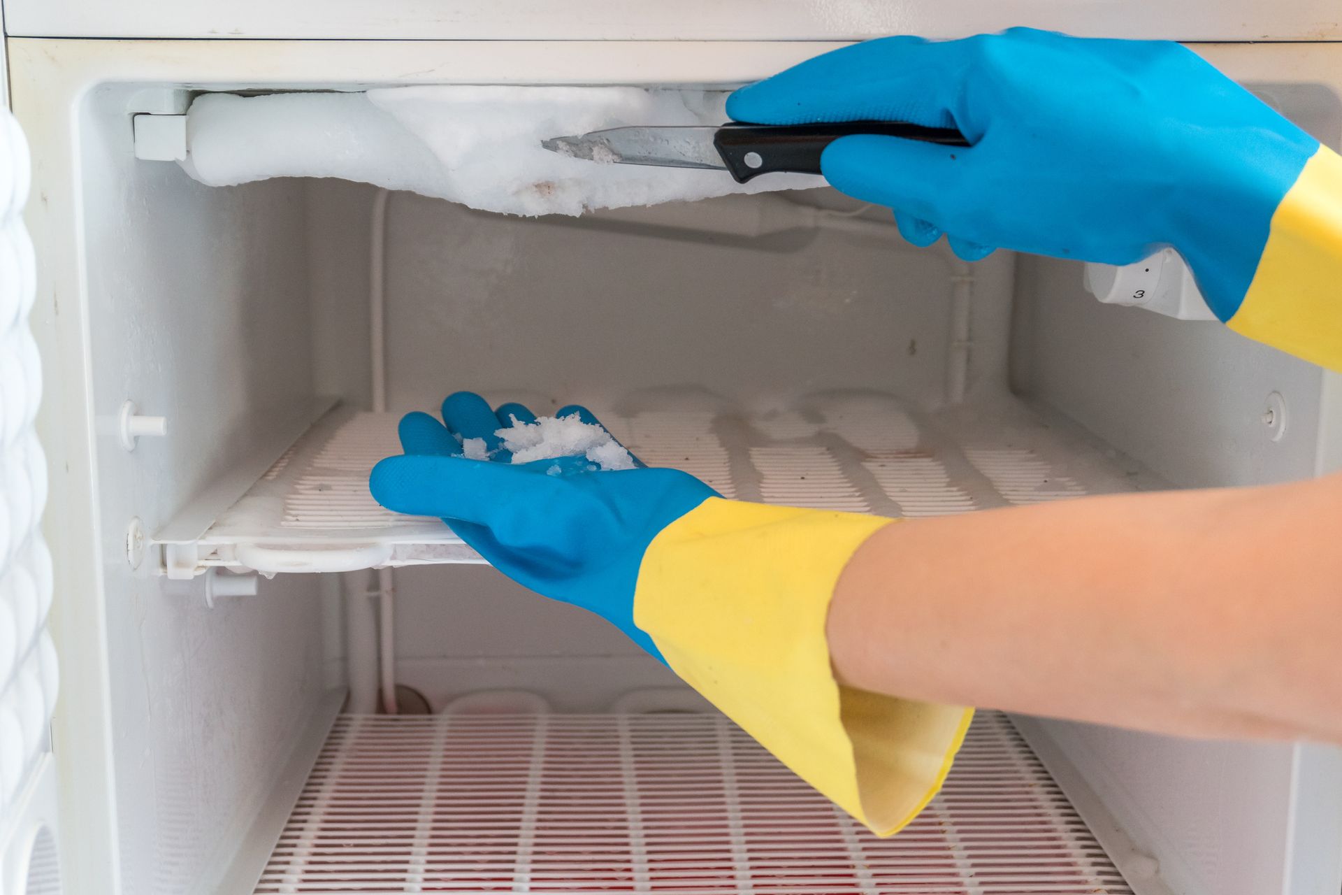 Person in blue and yellow gloves scraping ice from a freezer with a knife.