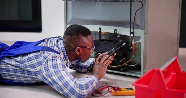Black man in overalls repairing a refrigerator on the floor with a flashlight and tools.