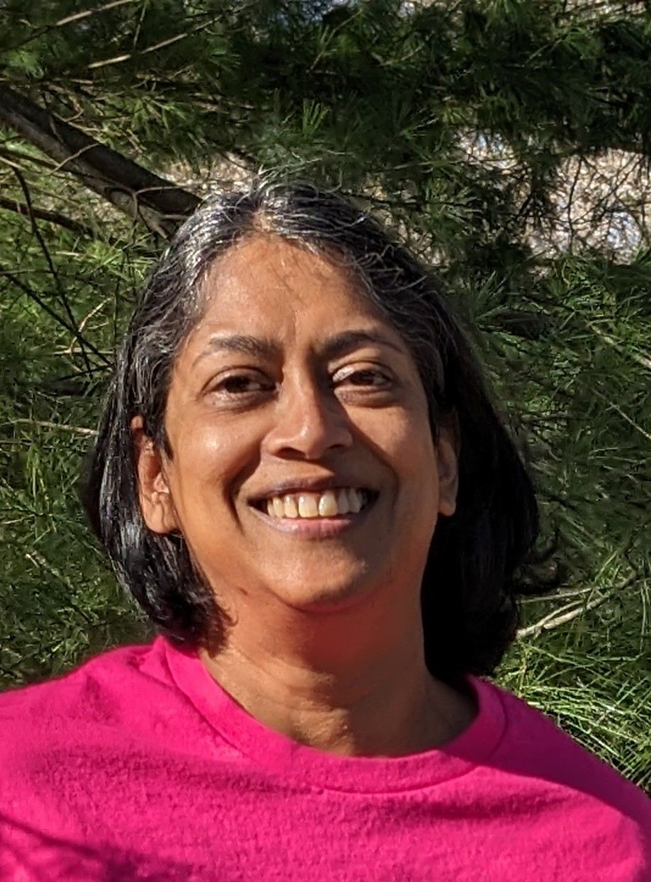 A woman in a pink shirt is smiling in front of a pine tree.