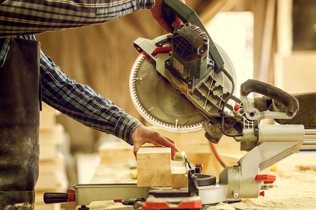 A man in a plaid shirt and work pants operating a motorized saw to cut a piece of wood.