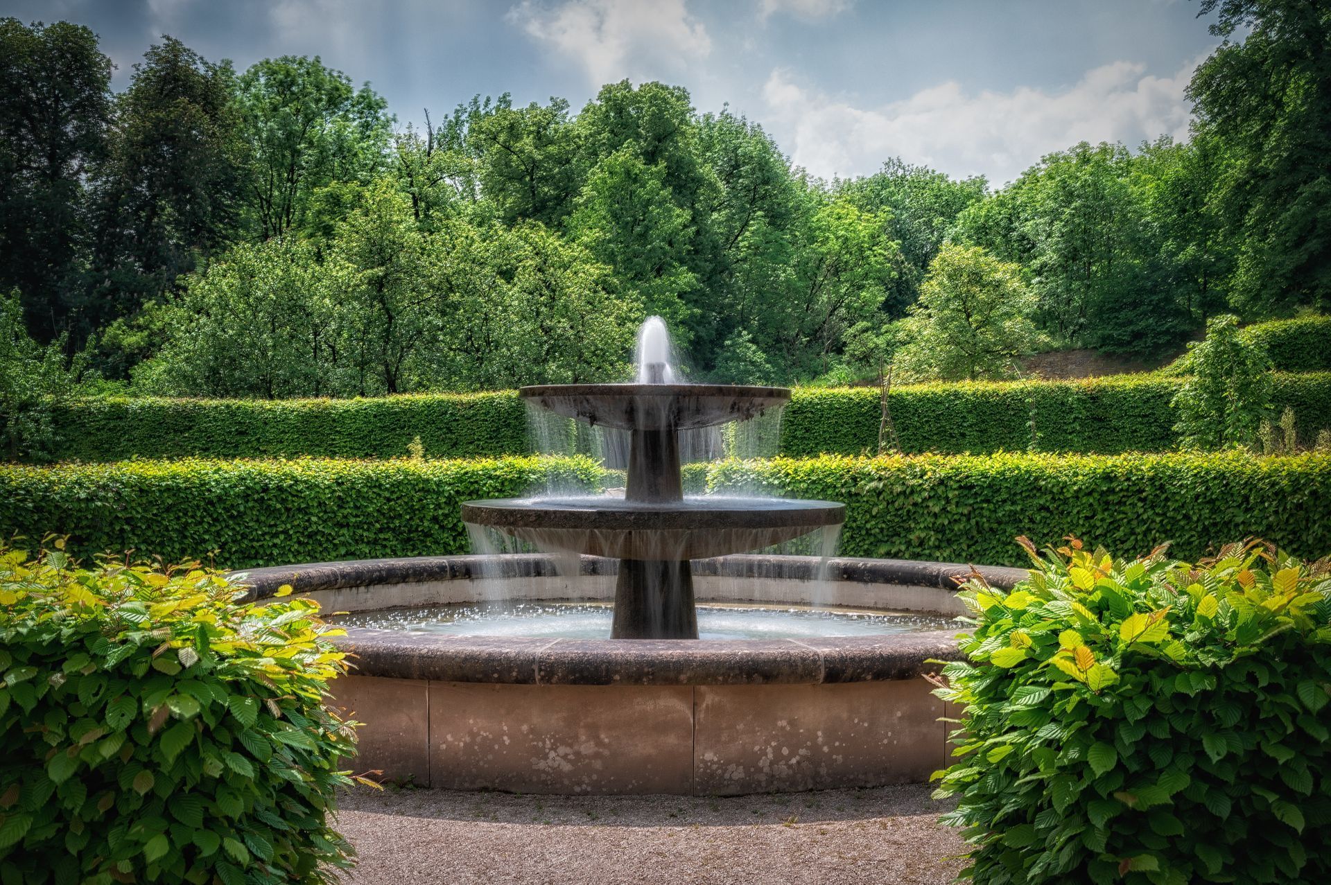 Serene outdoor scene featuring a decorative water fountain nestled amidst lush green hedges and towering trees.