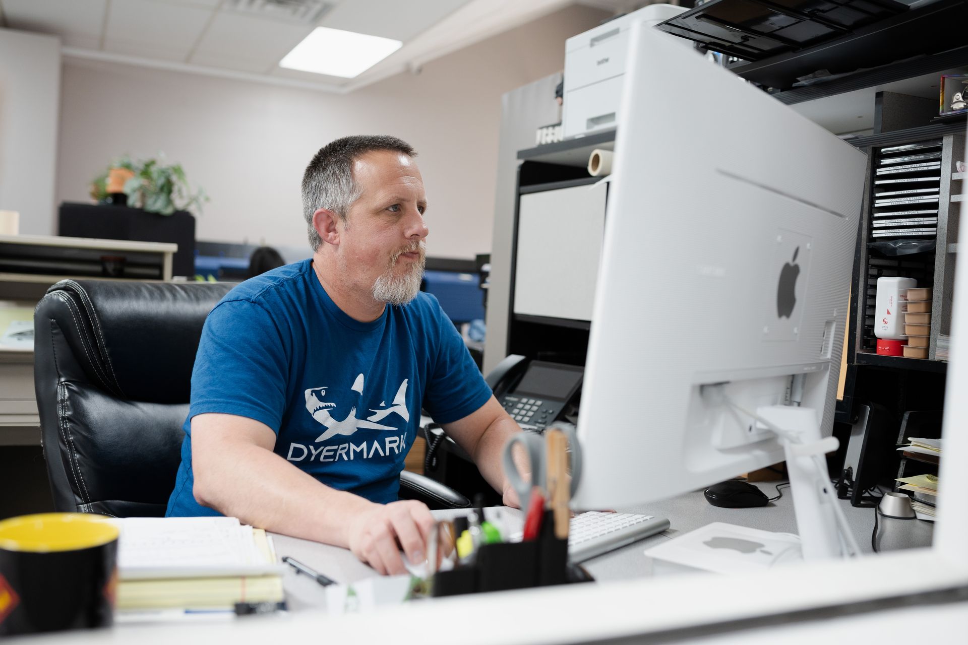 A man in a blue shirt is sitting at a desk in front of a computer.