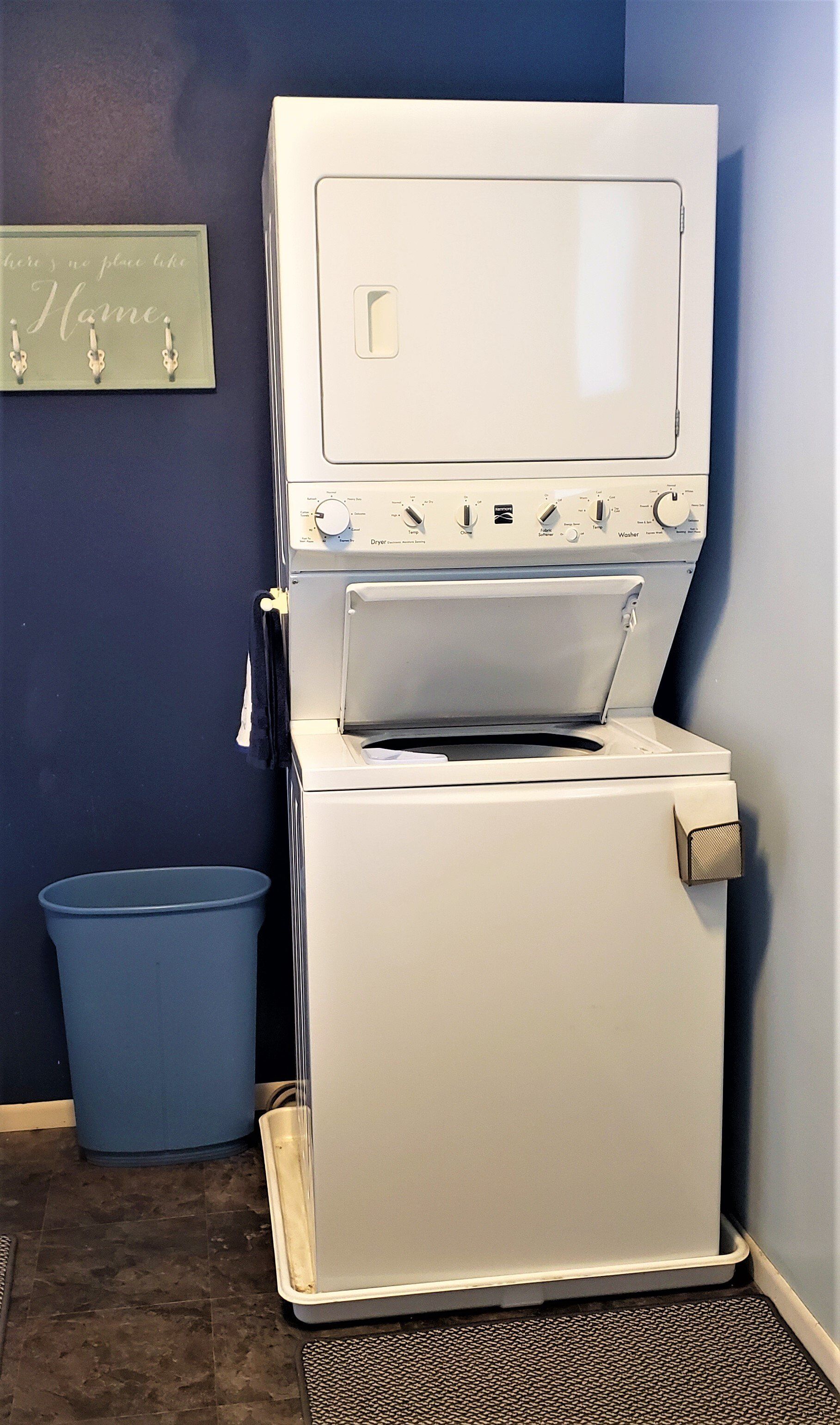 Stackable white washer and dryer in a corner next to a blue trash can and a blue wall.