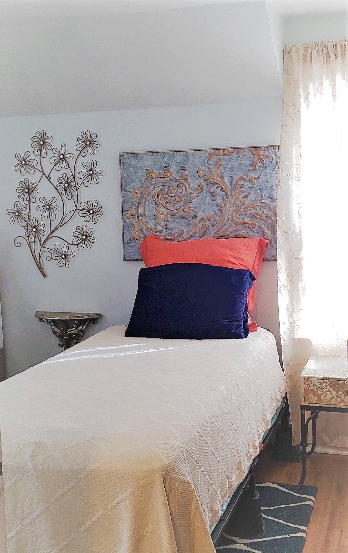 Bed with pillows against a wall, art on either side. A sheer curtain is on the right.