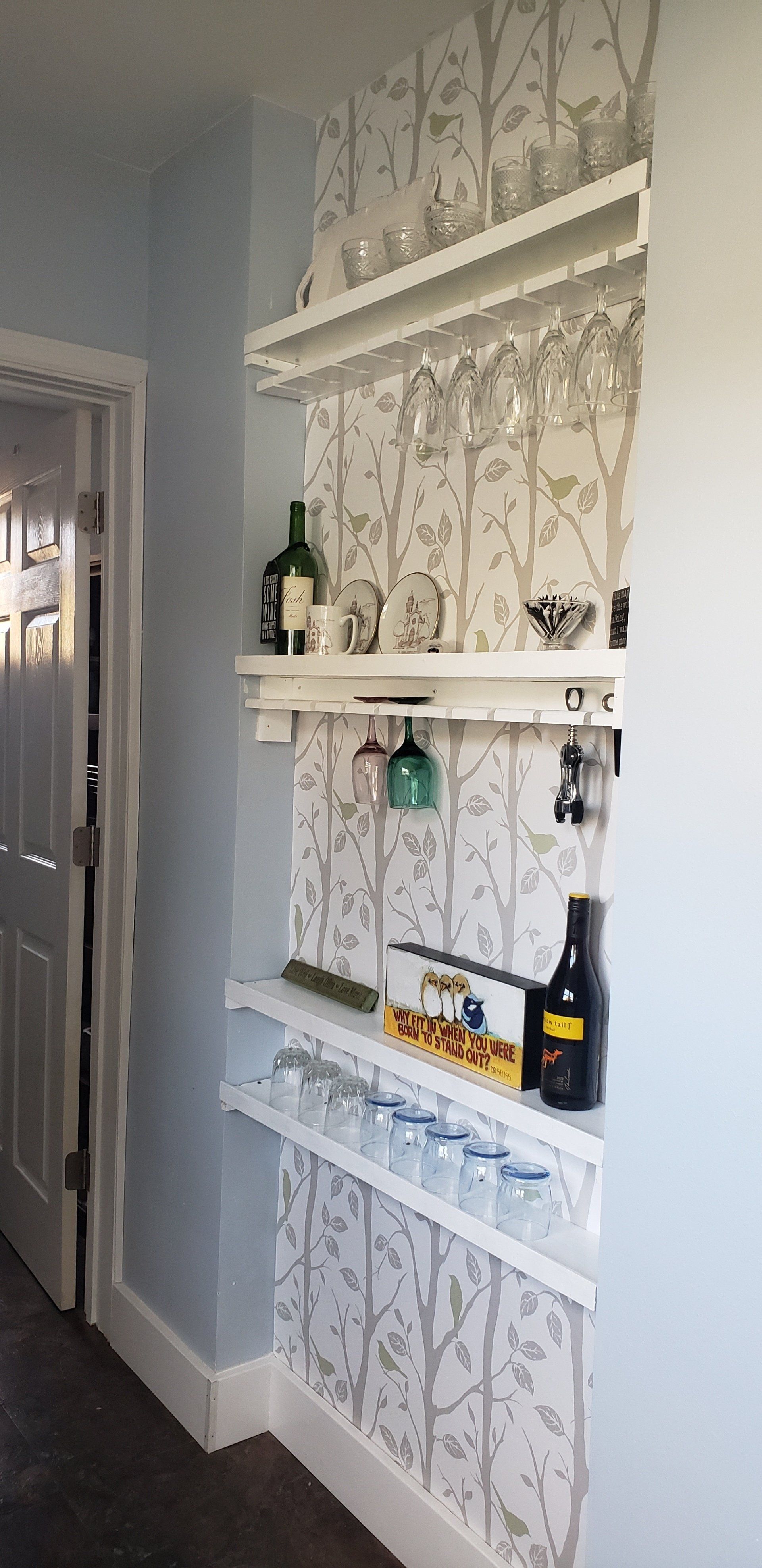 Built-in white bar shelves on a light gray wall with patterned wallpaper, holding glasses and bottles.