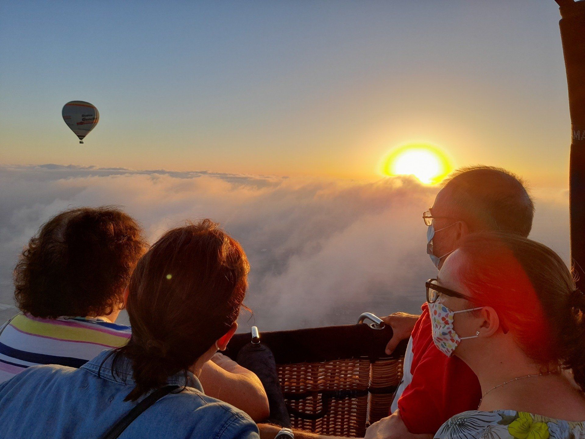 Un grupo de personas observa un globo aerostático volando sobre las nubes al atardecer.