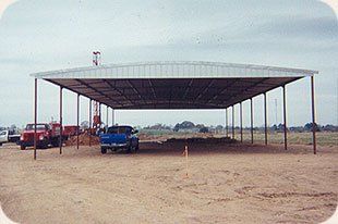 A blue truck is parked under a metal structure in a dirt field.