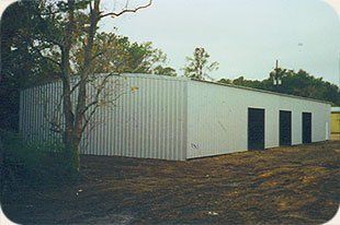A large white building with black doors is sitting in the middle of a dirt field.