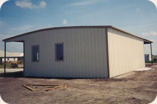 A white metal building with a porch is sitting in the middle of a dirt field.