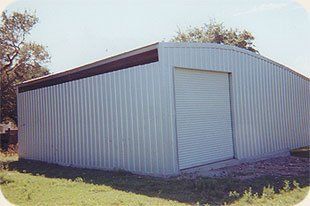 A large metal building with a roller door is sitting in the middle of a grassy field.