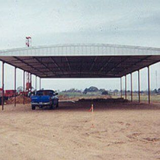 A blue truck is parked under a roof in a dirt field.