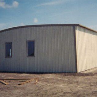 A white metal building with three windows is sitting in the middle of a dirt field.