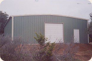 A green building with a white garage door is surrounded by bushes.