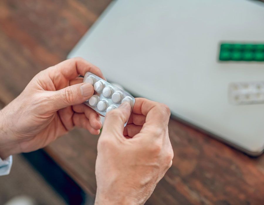Hands holding a blister pack of white pills over a desk with a laptop in the background