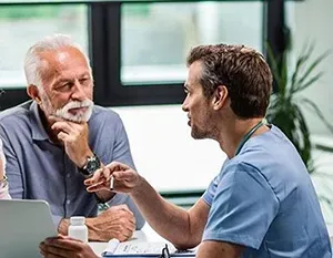 Two people in blue scrubs talking at a table in a bright office, one holding a clipboard.