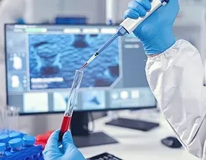 Gloved hands pipetting red liquid in a lab, with a medical scan displayed on a computer monitor.