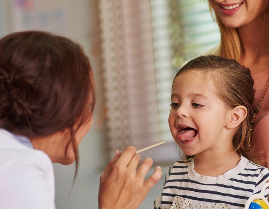 Adult feeding a smiling child with a spoon while another person stands nearby indoors