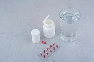 Medicine bottle, capsule, blister pack, and glass of water on a gray surface