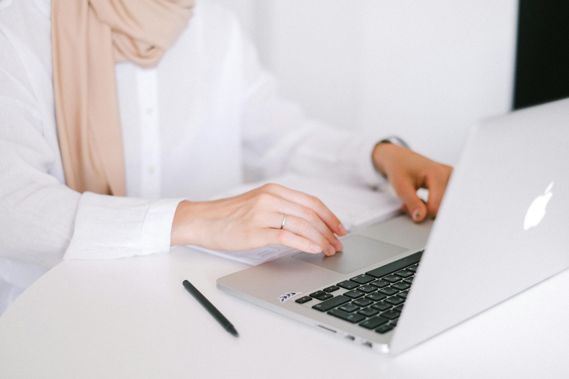 Person typing on a laptop at a white desk, with a pen beside it and a beige scarf visible.