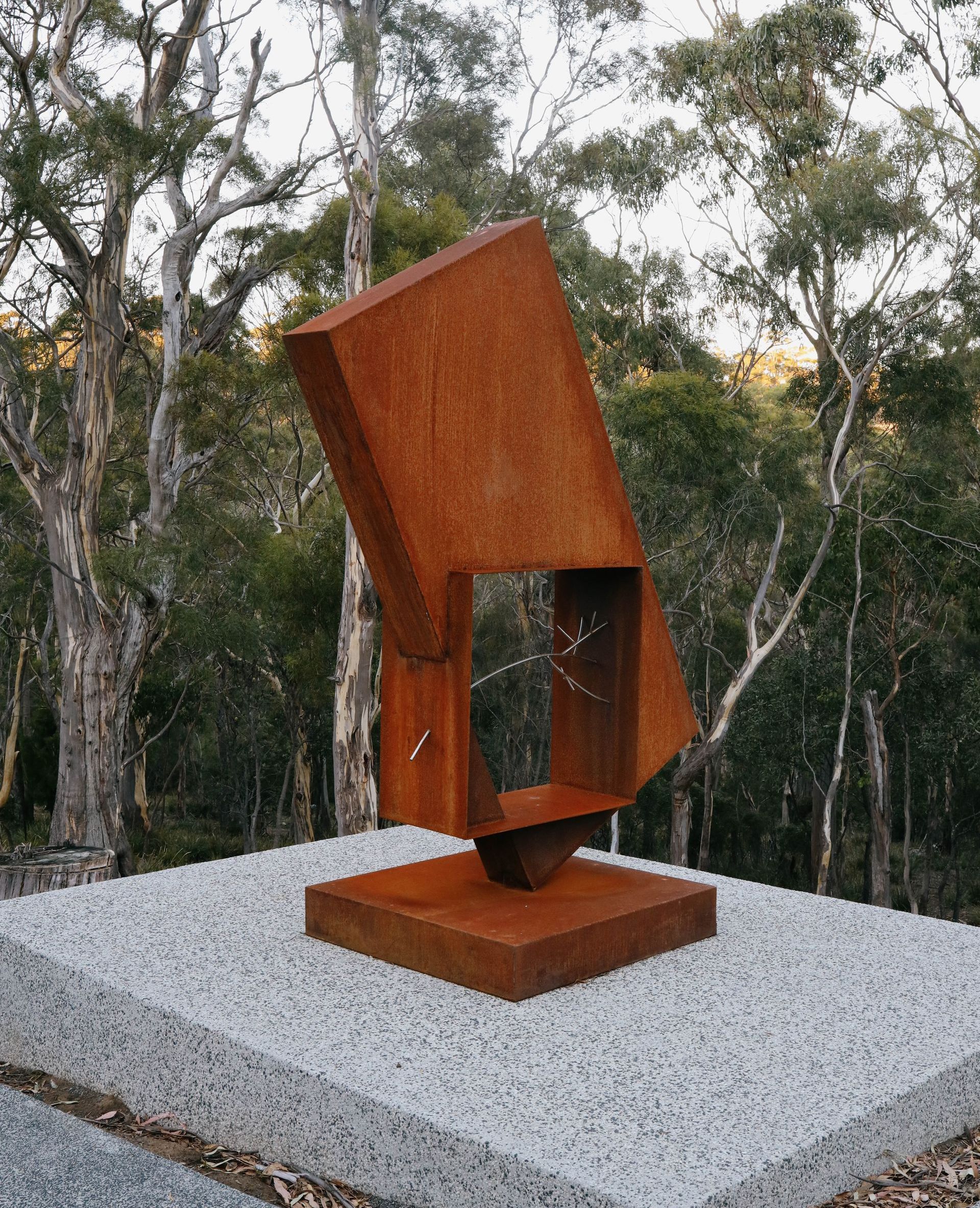 A rusty sculpture is sitting on top of a gravel platform in the middle of a forest.