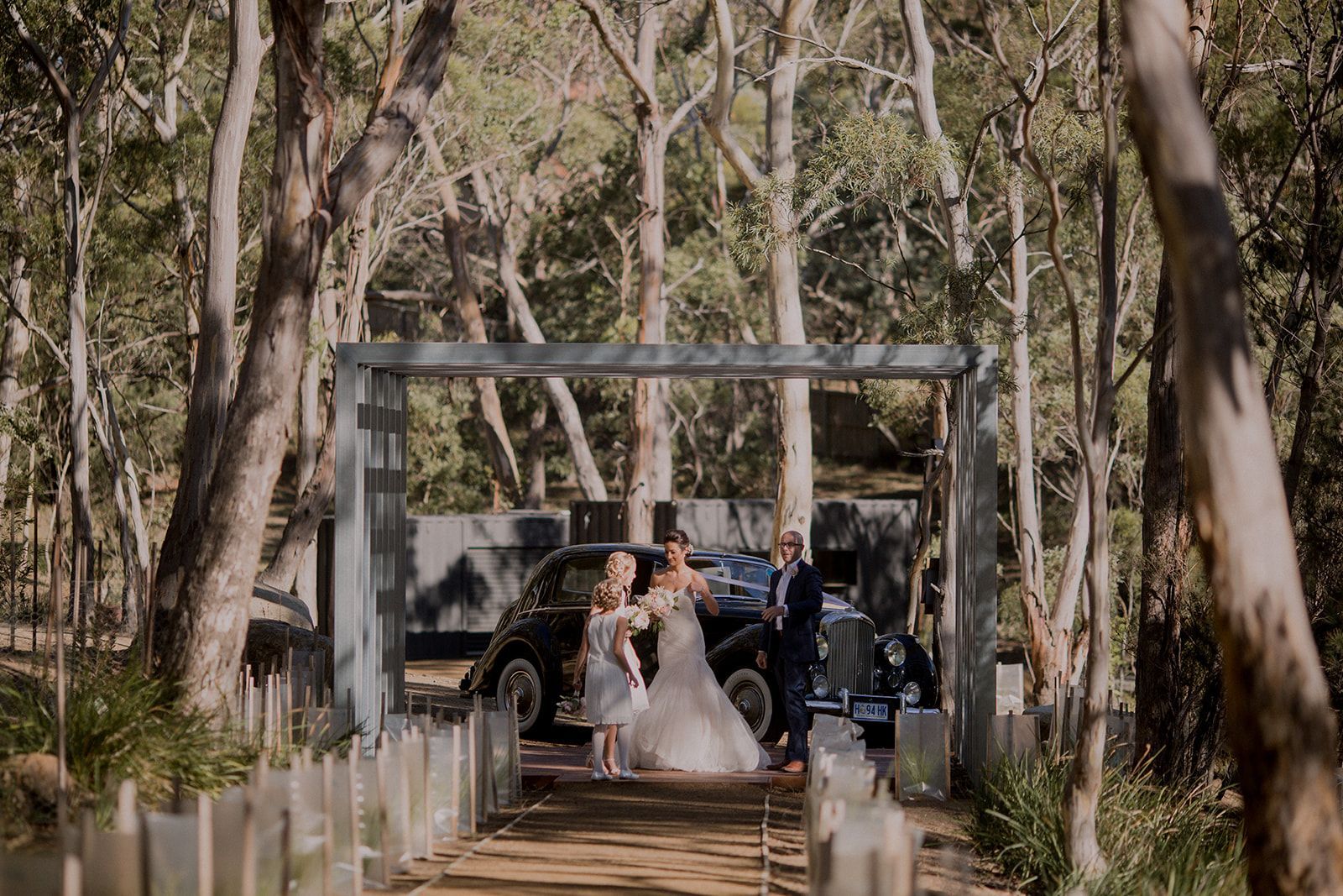 A bride and groom are standing next to a car in the woods.
