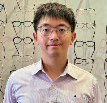 Joe Wang with dark hair and glasses poses in front of a wall display of eyeglass frames.