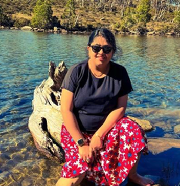 Payel wearing dark sunglasses and a floral skirt sits on a large driftwood log in a clear, sunlit alpine lake.