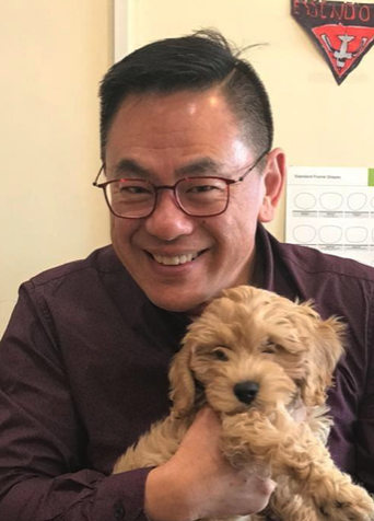Ning wearing glasses and a purple shirt smiling while holding a small, fluffy, light-brown puppy.