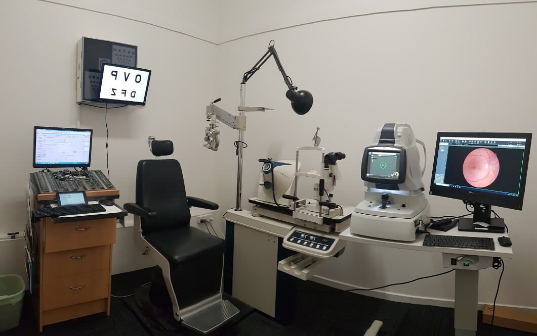 An eye exam room with a patient chair, phoropter, slit lamp, and multiple monitors displaying images of an eye.