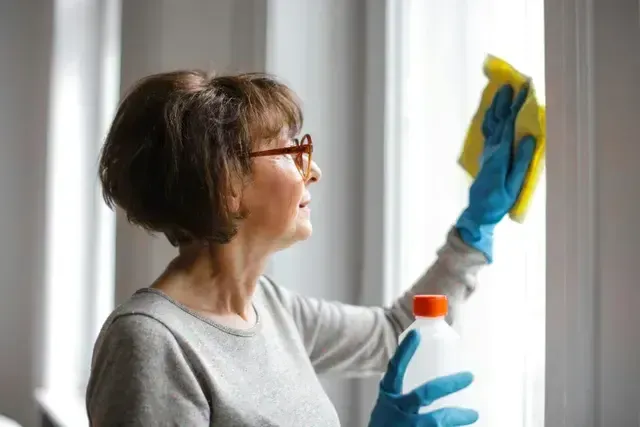 Woman wearing glasses and gloves, cleaning a window with a yellow cloth.