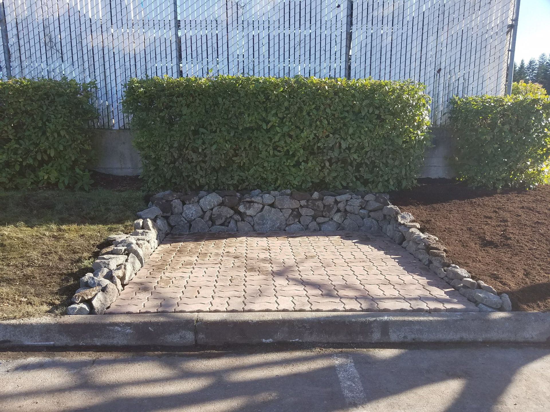 Brick paved area with stone border, backed by greenery and a white slatted fence.