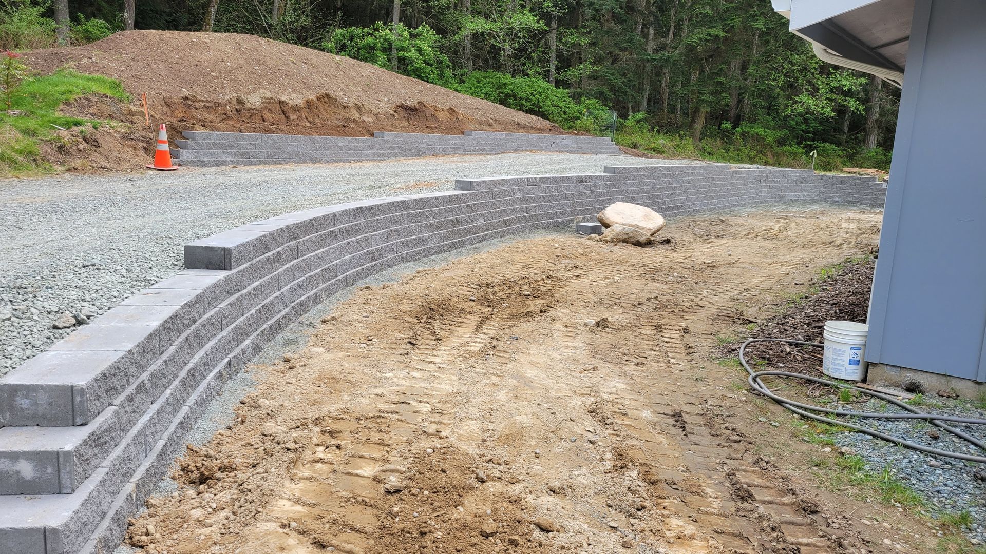 Curved retaining wall built from gray blocks with gravel driveway, dirt, and a forest background.