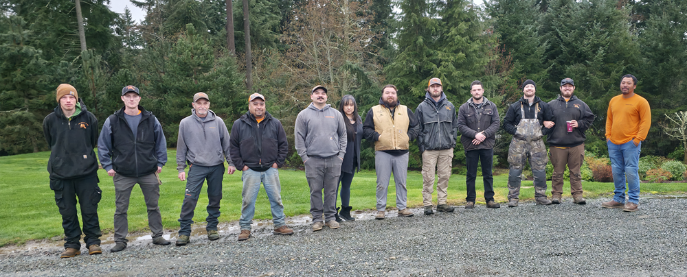 Landscaping crew poses outdoors with orange shirts and mowers.