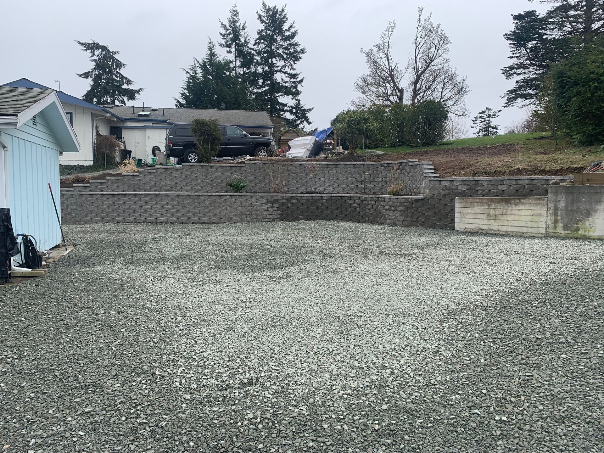 Gravel driveway in front of tiered retaining walls made of gray blocks. A house and trees are in the background.