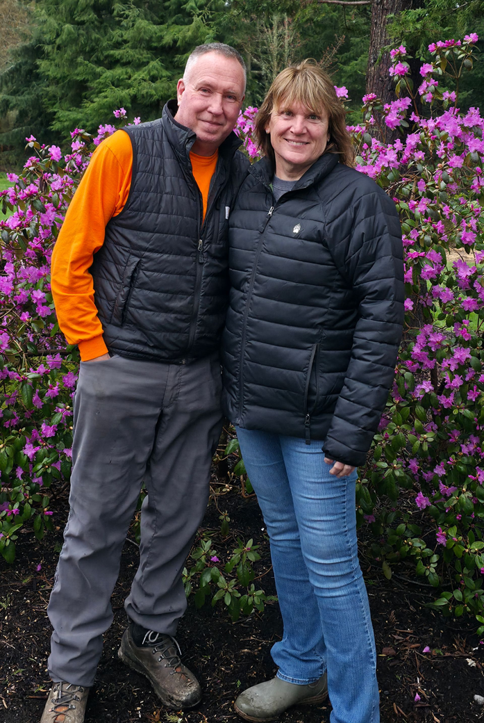 Group of people in orange shirts with lawn mowers; posing for a picture in a grassy area with trees.