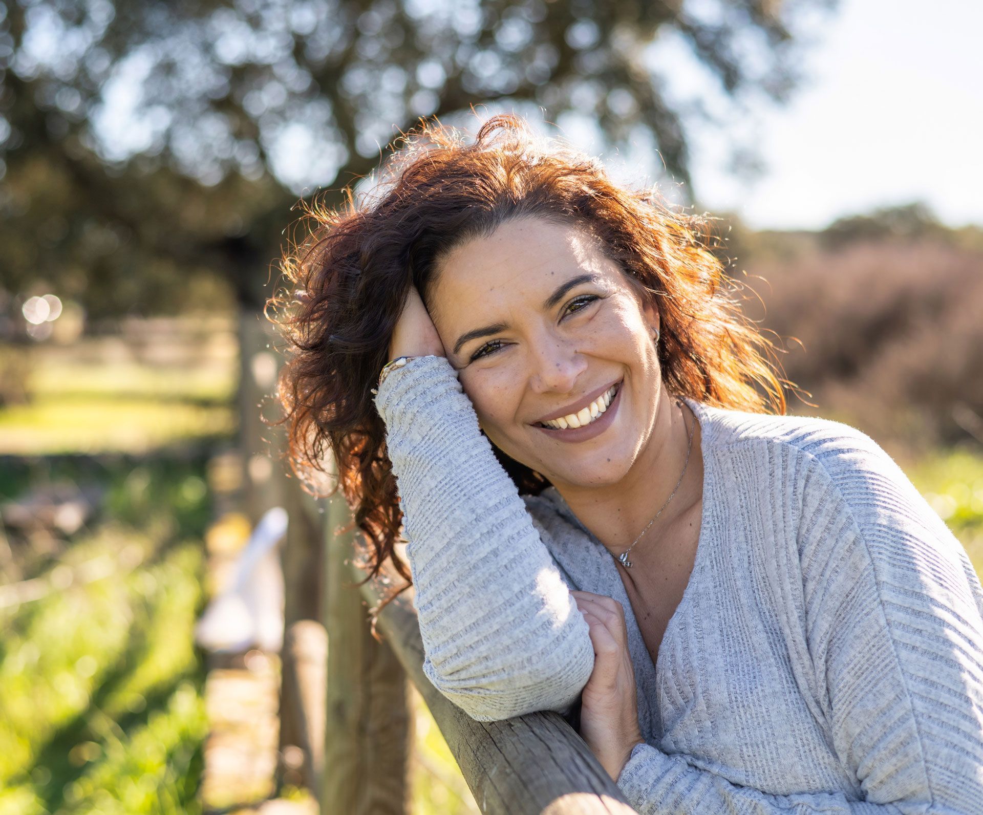 Woman with curly auburn hair smiles, resting on a wooden fence outdoors in sunlight.