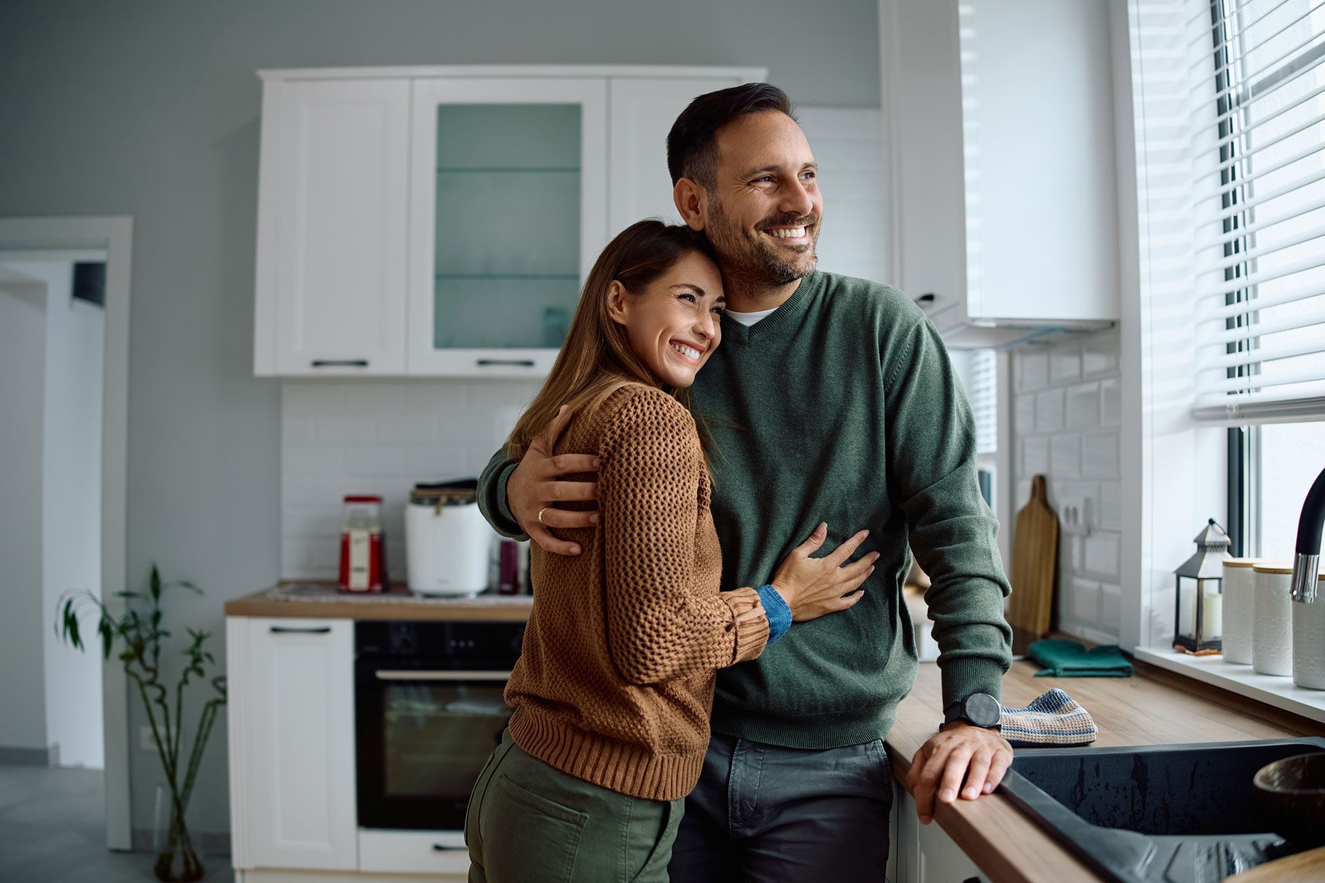 Couple hugging in a kitchen, looking out the window, smiling.