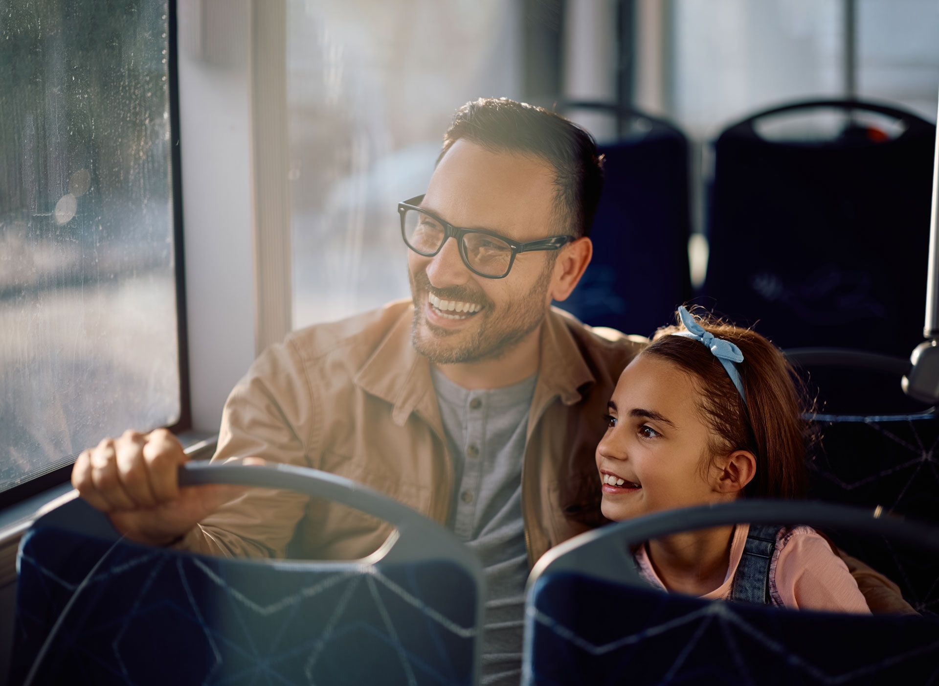 Father and daughter smiling on a bus, looking out window. Sunlight shines on them.