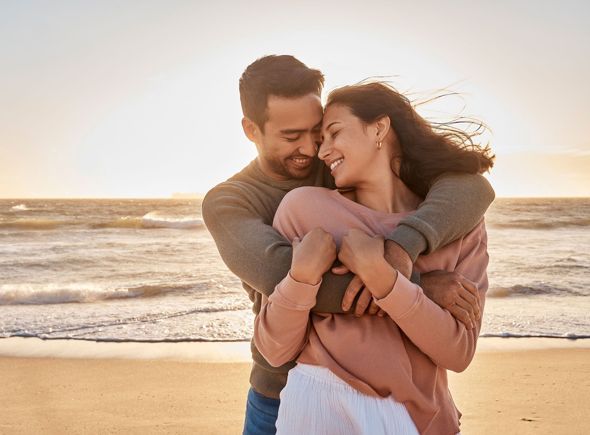 Couple hugging on a beach at sunset, smiling. Ocean waves, golden light.