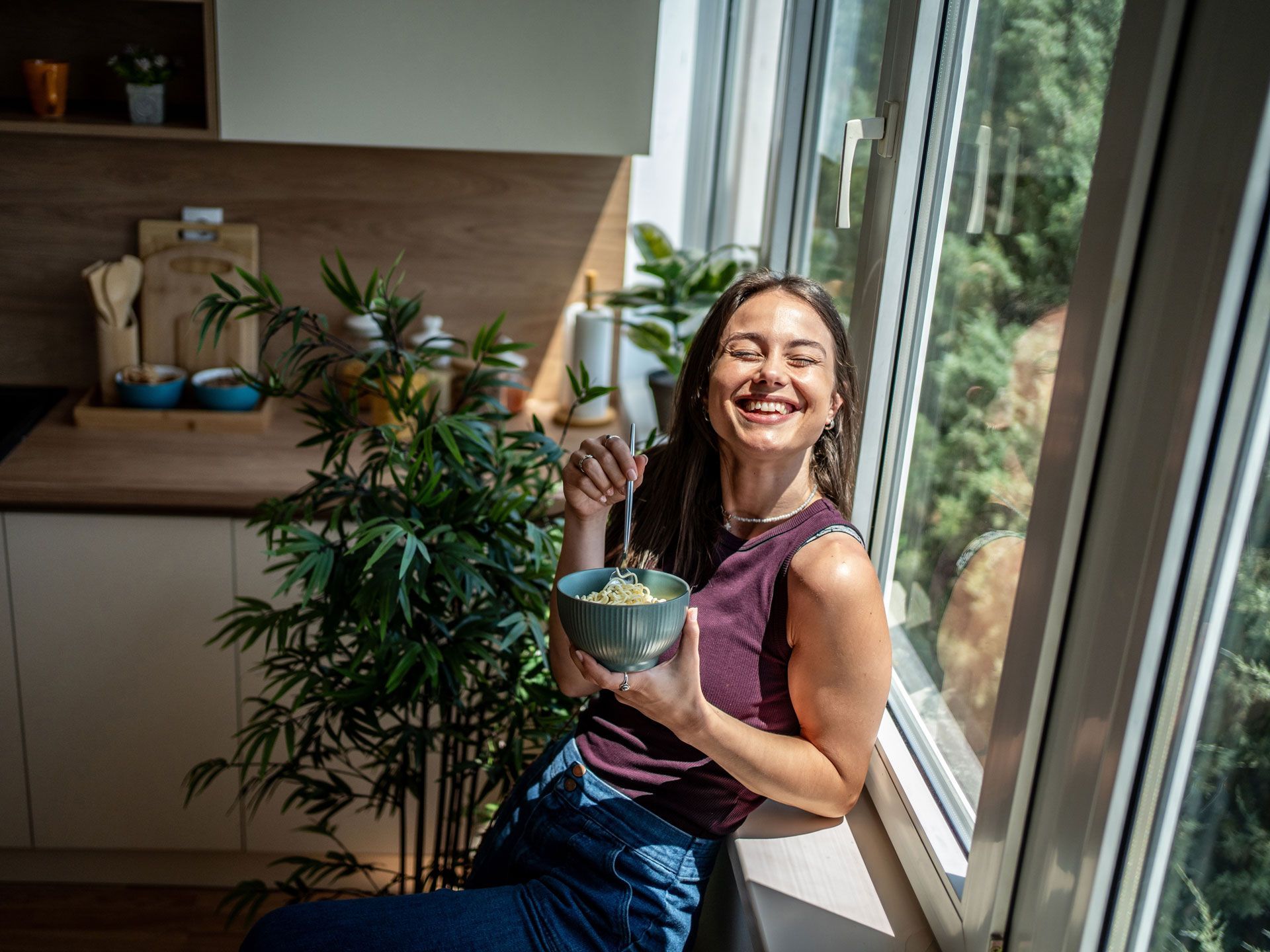 Woman smiles, sitting by a window, holding a bowl, enjoying a meal in her kitchen.
