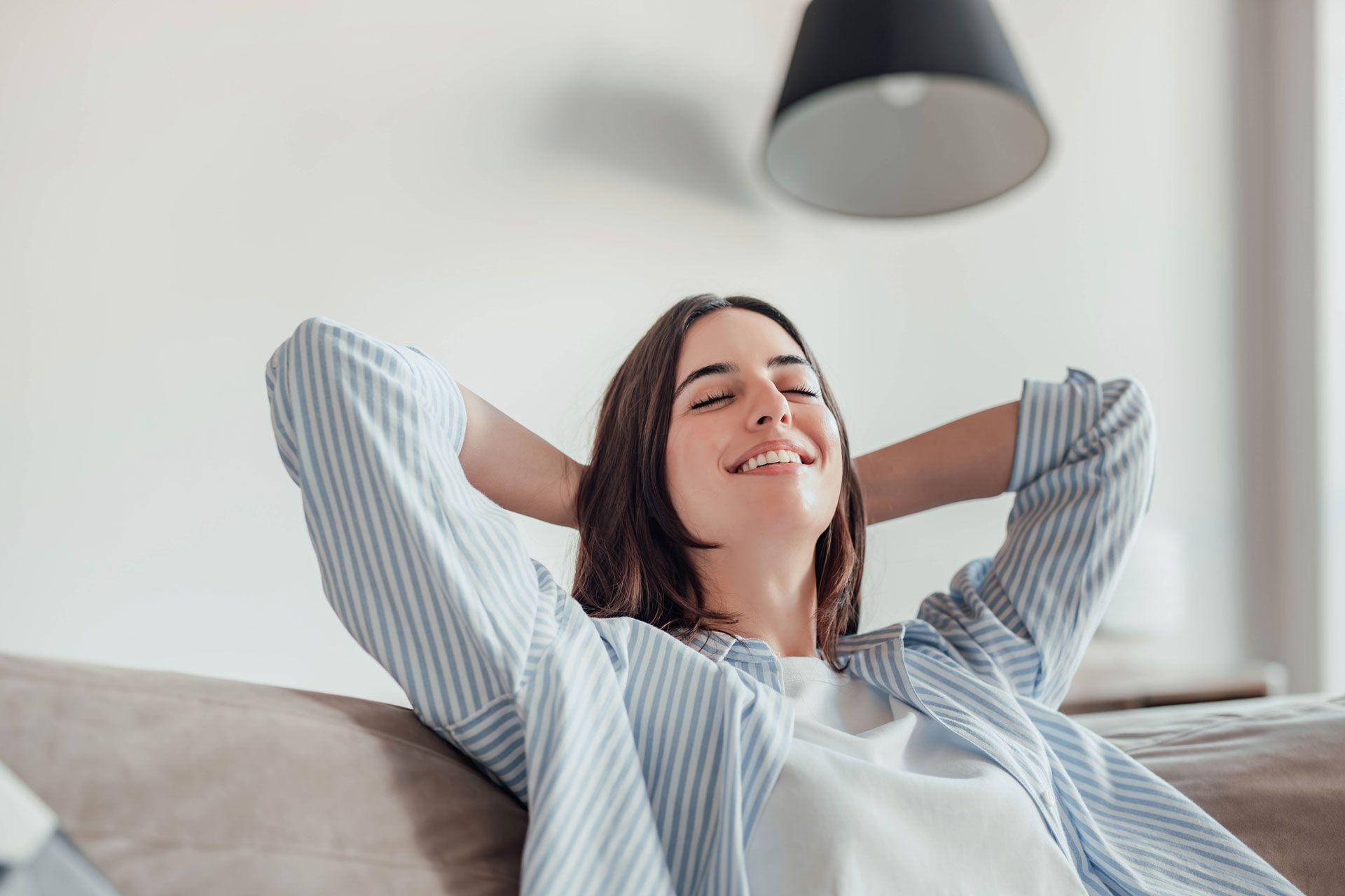 Woman relaxing on a sofa, smiling with hands behind her head, indoors with overhead light.