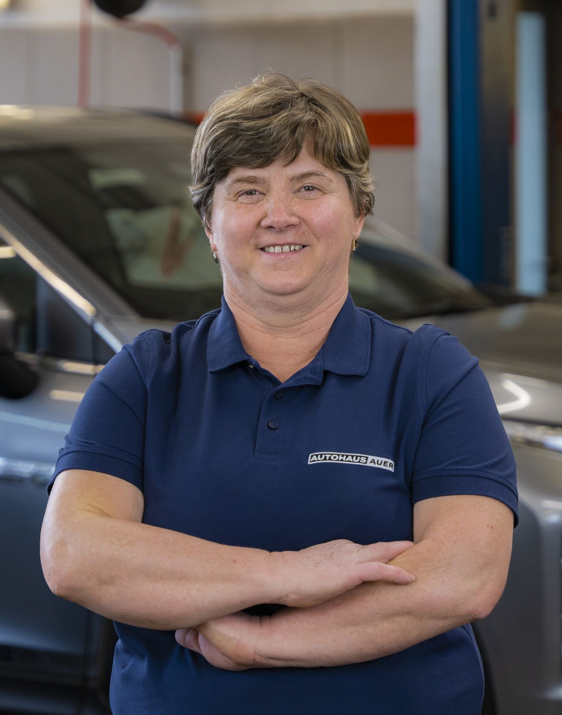 Eine Frau in einem blauen Hemd steht mit verschränkten Armen vor einem Auto.