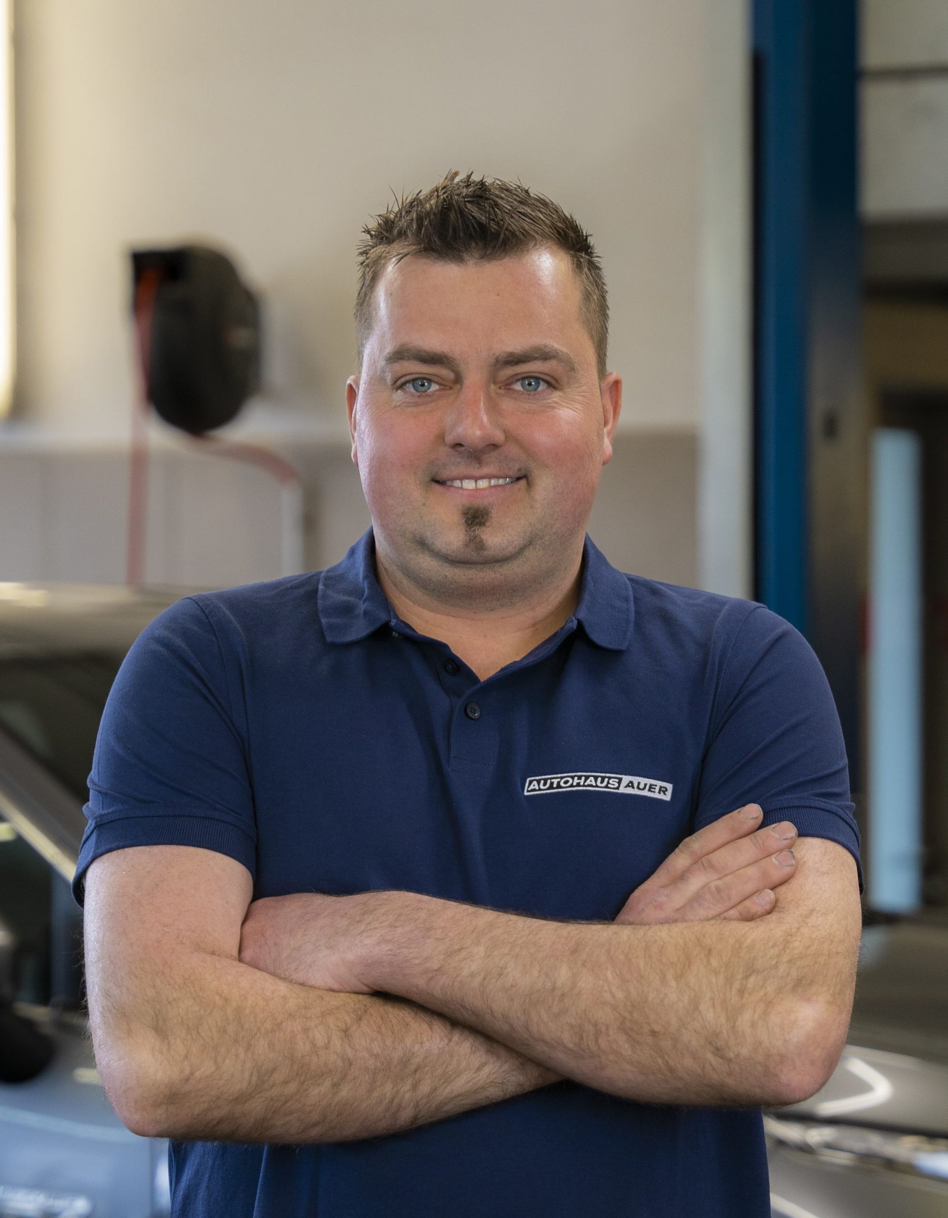 Ein Mann in einem blauen Hemd steht mit verschränkten Armen vor einem Auto.