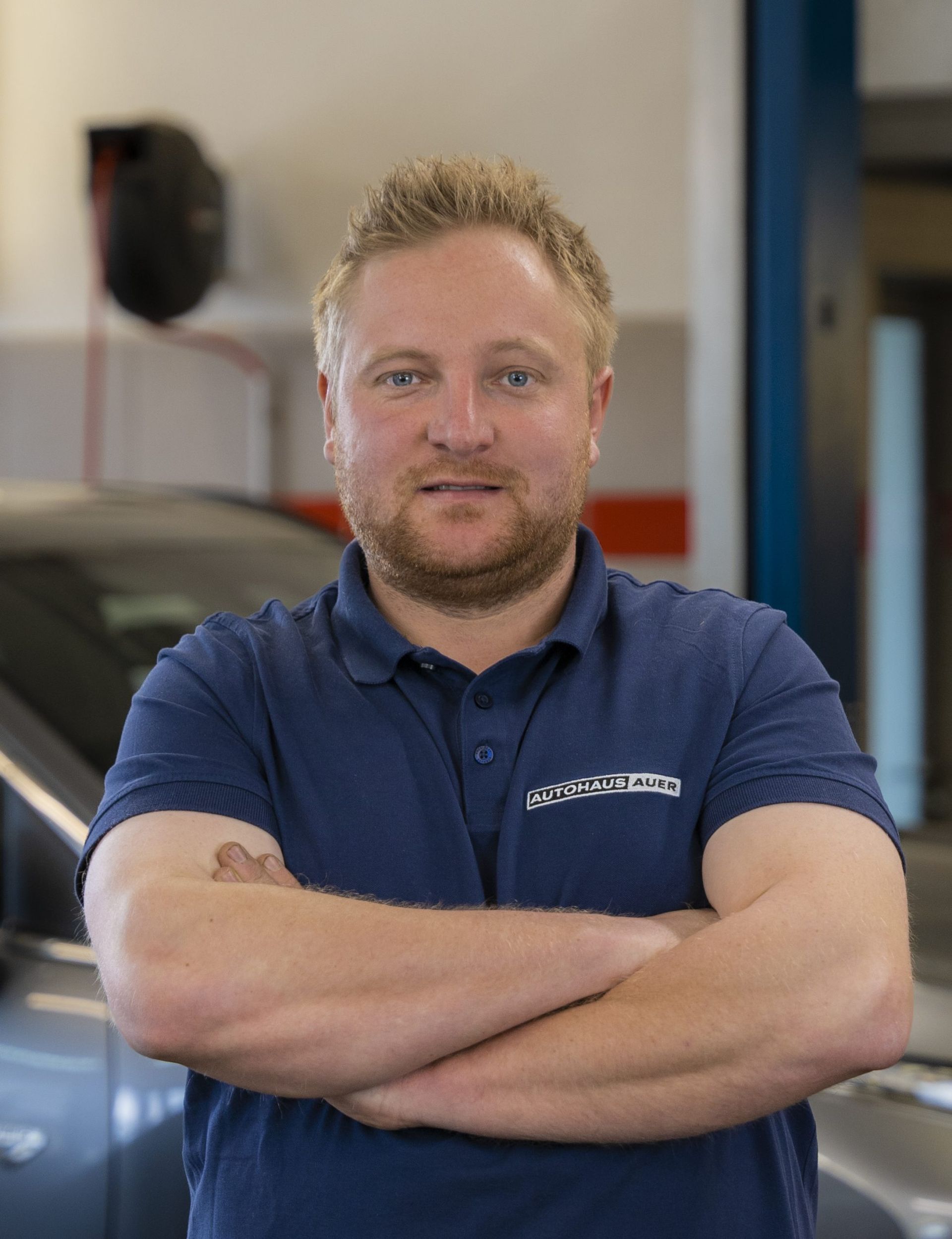 Ein Mann in einem blauen Hemd steht mit verschränkten Armen vor einem Auto.