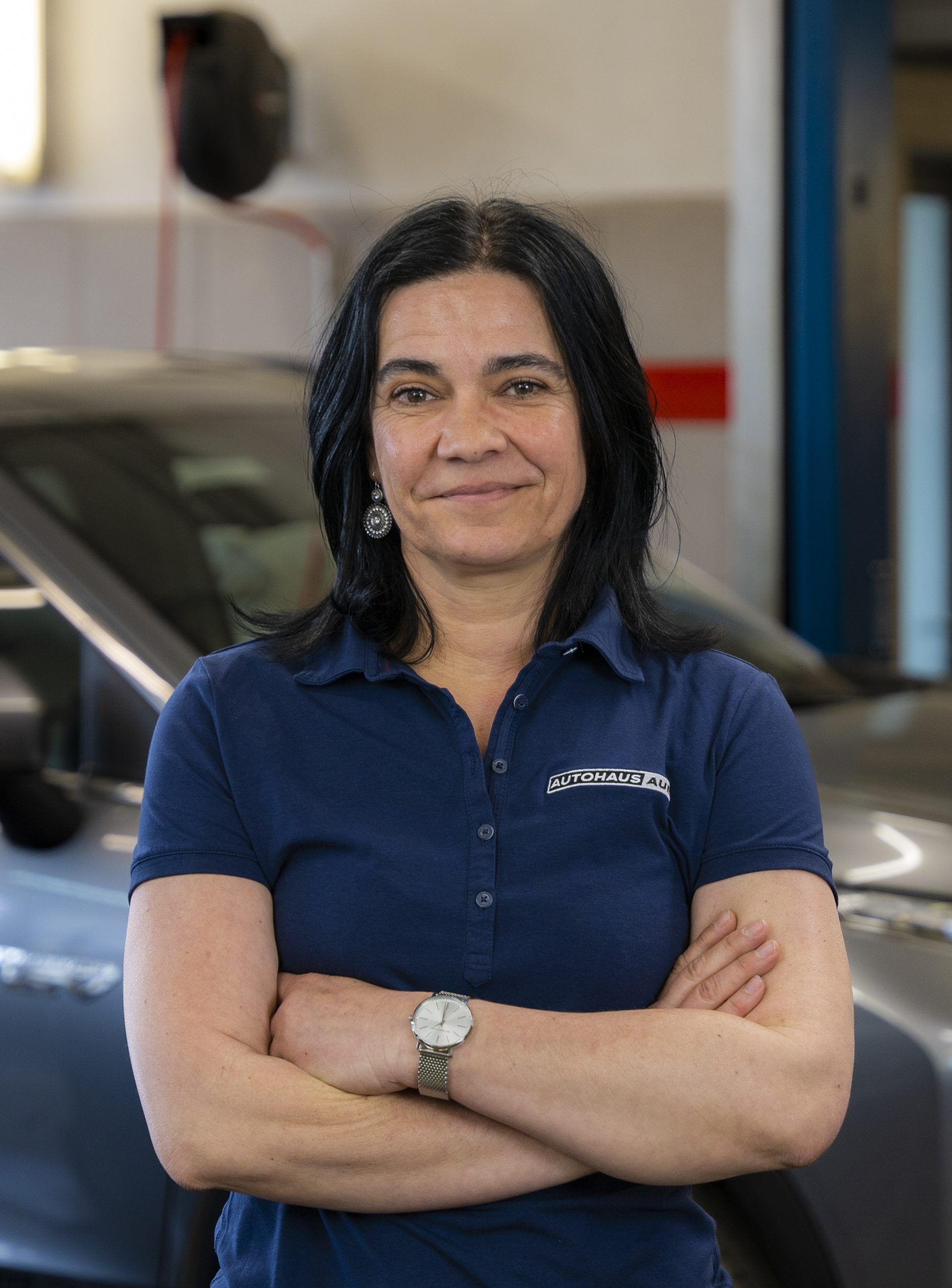 Eine Frau in einem blauen Hemd steht mit verschränkten Armen vor einem Auto.