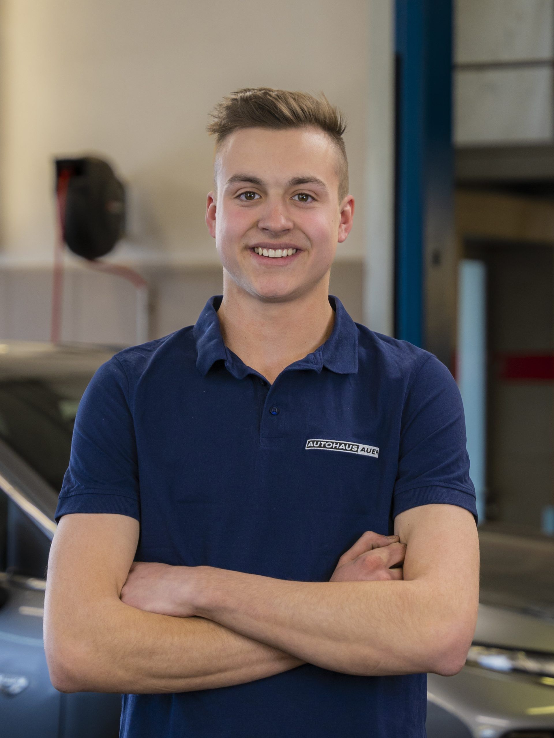 Ein junger Mann in einem blauen Hemd steht mit verschränkten Armen vor einem Auto.