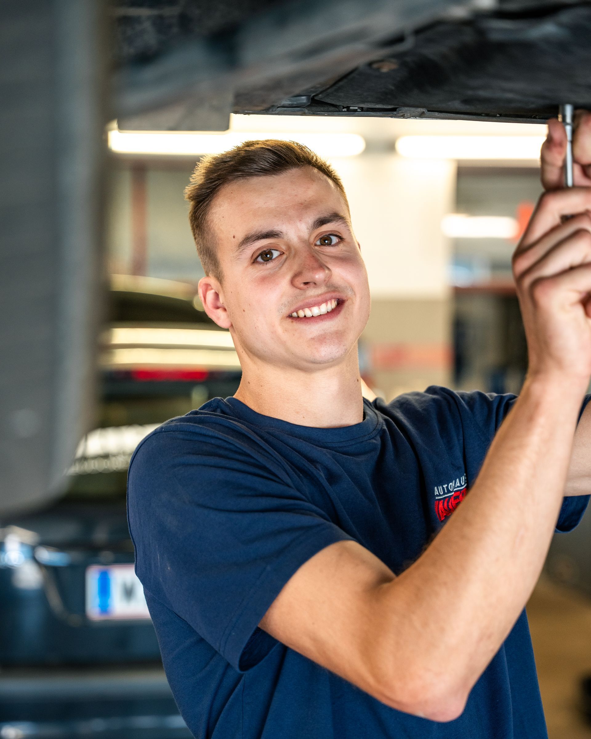 Ein junger Mann in einem blauen Hemd steht mit verschränkten Armen vor einem Auto.
