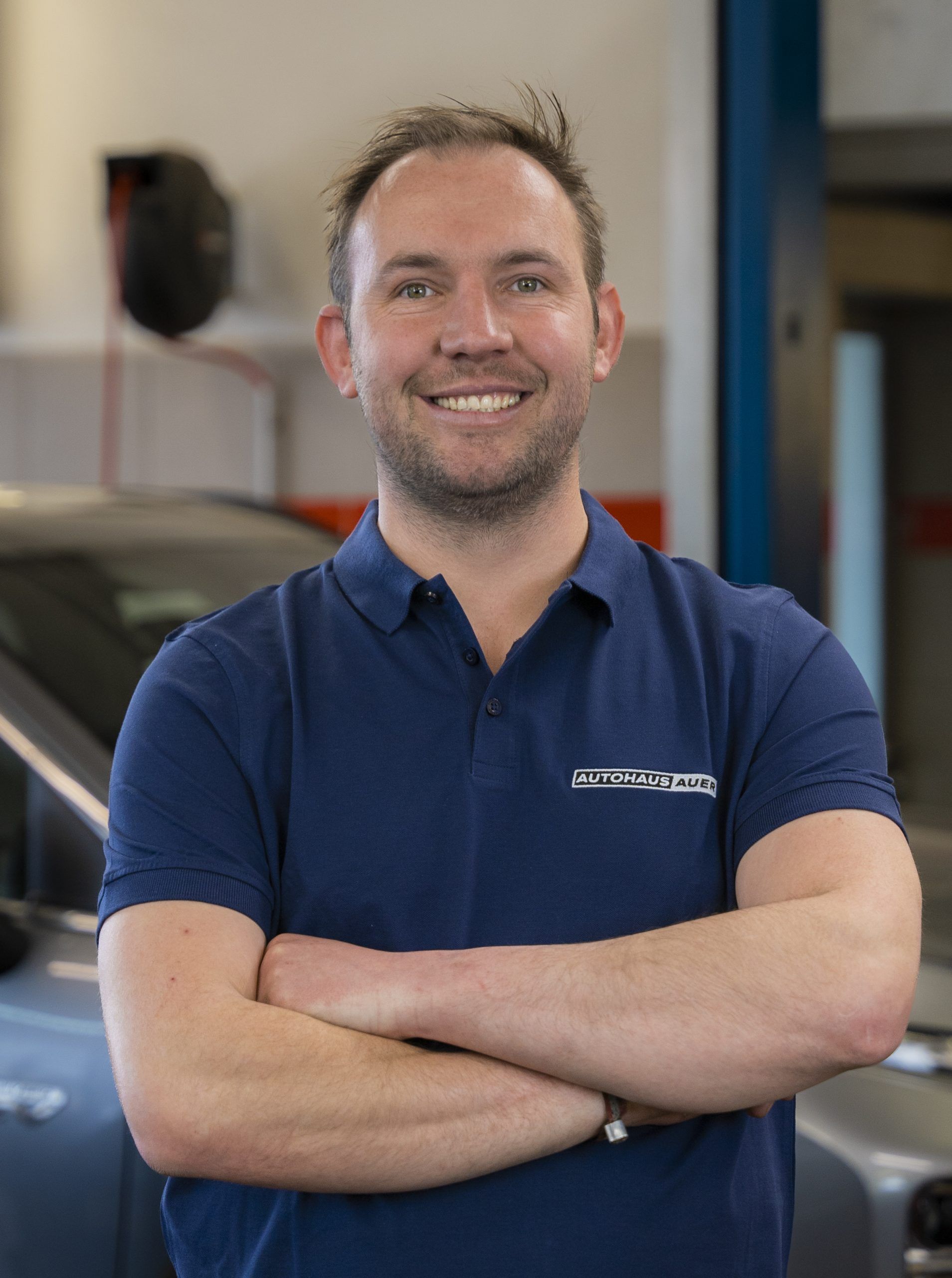 Ein junger Mann in einem blauen Hemd steht mit verschränkten Armen vor einem Auto.