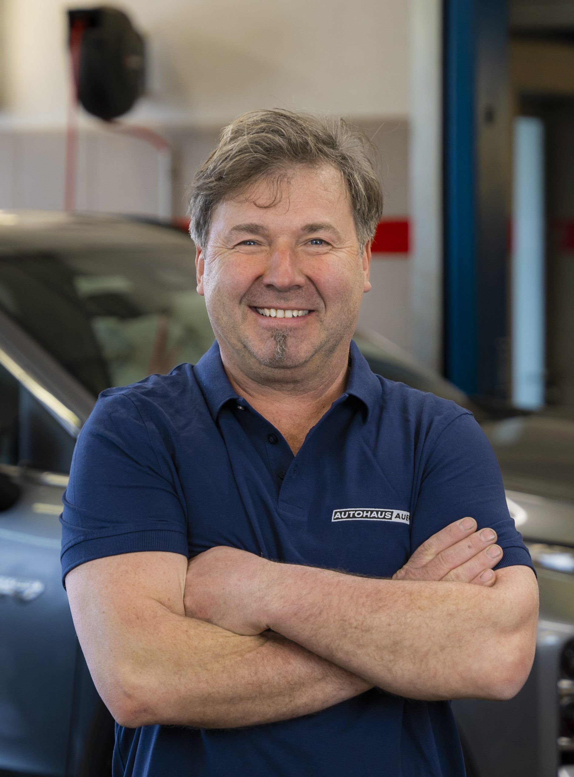 Ein Mann in einem blauen Hemd steht mit verschränkten Armen vor einem Auto.